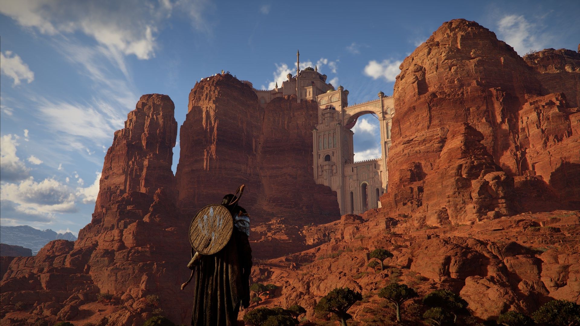 Standing in a desert with a wooden, circular shield on his back, the hero looks toward red stone cliffs with a stone gate rising between them.