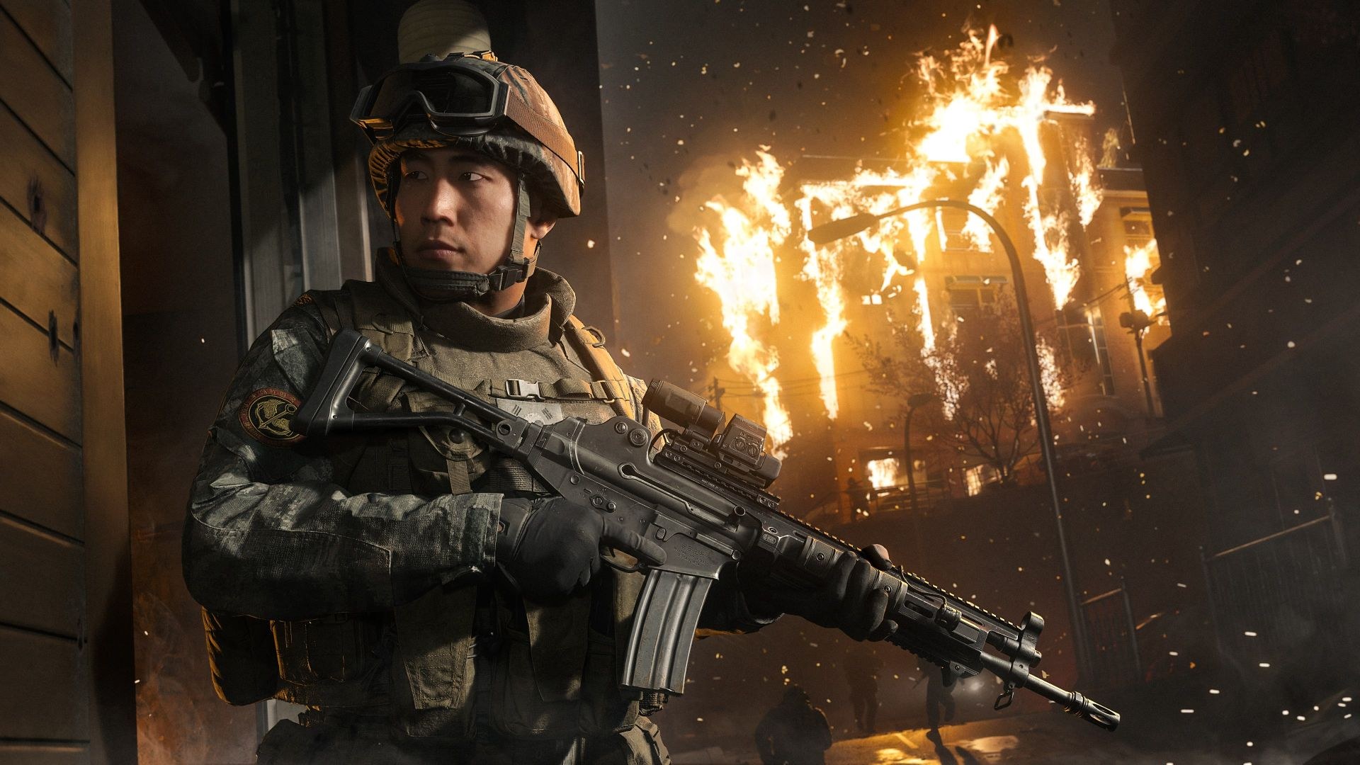A closeup view of a soldier looking off to his right, while a multi-level building goes up in flames behind him and people run through the street.