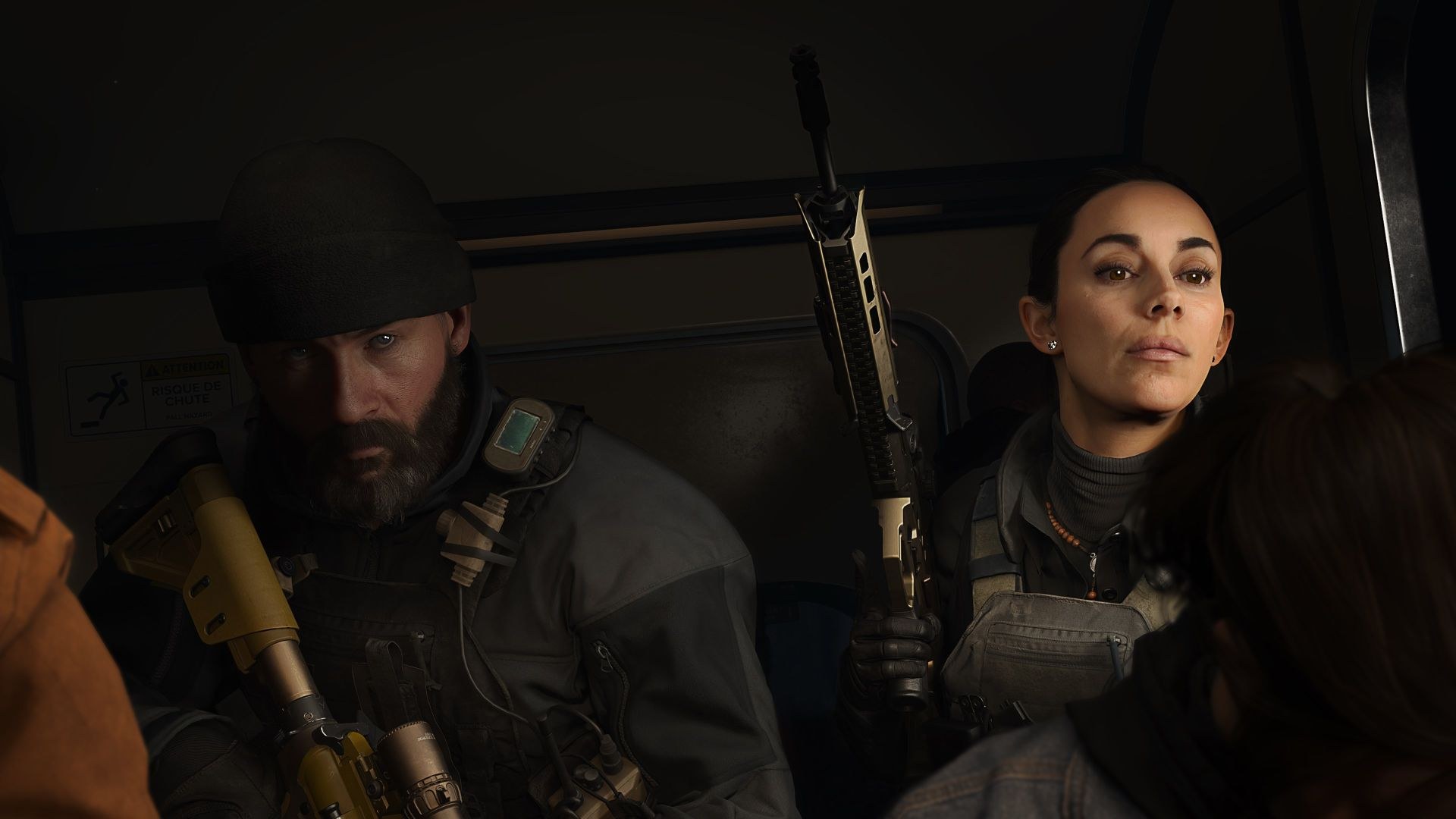 A bearded soldier in a beanie sits next to a woman with her gun held pointed toward the ceiling, both inside a vehicle.