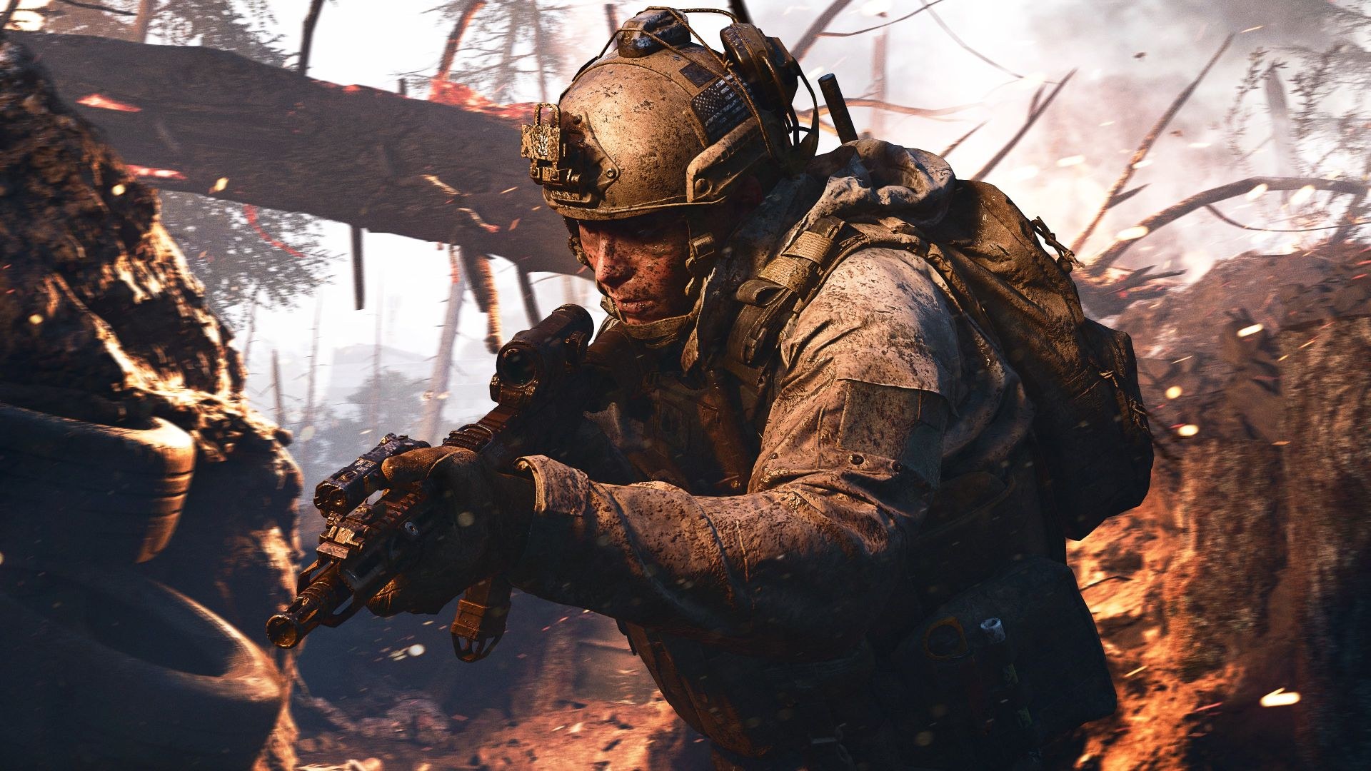 A soldier in dirty gear holds a rifle pointed slightly downward as he advances through a trench with a fallen tree lying across it in the background.